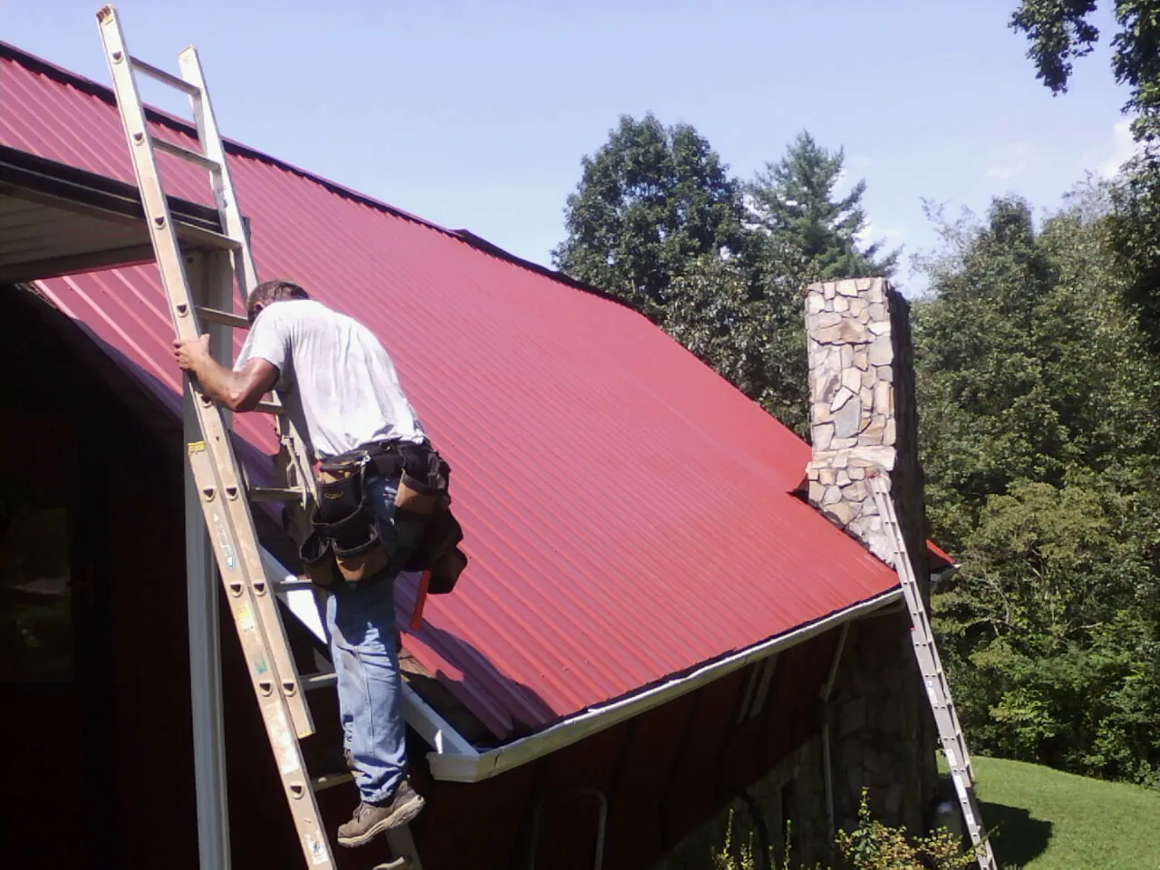 Roofer on a ladder beside a red metal roof and stone chimney — Soffit & Fascia Repair by Roofing Brothers in Osburn